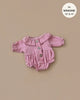 The Leonore Ruffled Jumpsuit in Tea Pink for Minikane dolls is a pink, long-sleeve button-up romper with ruffled cuffs and a round collar made of soft cotton, displayed on a beige background and fits 13" to 14" sizes.