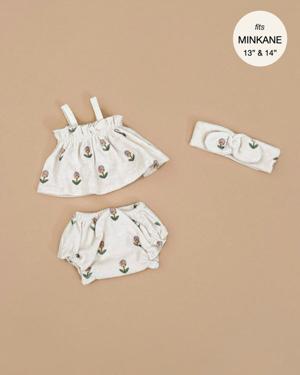 The Minikane Doll Clothing "Mila Soft Flowers Set In Terrycloth" by Emile & Ida is a light-colored set on a beige background, featuring a sleeveless top, bloomers, and floral headband. Designed in France, the hand-washable set fits Minikane 13” & 14” dolls.