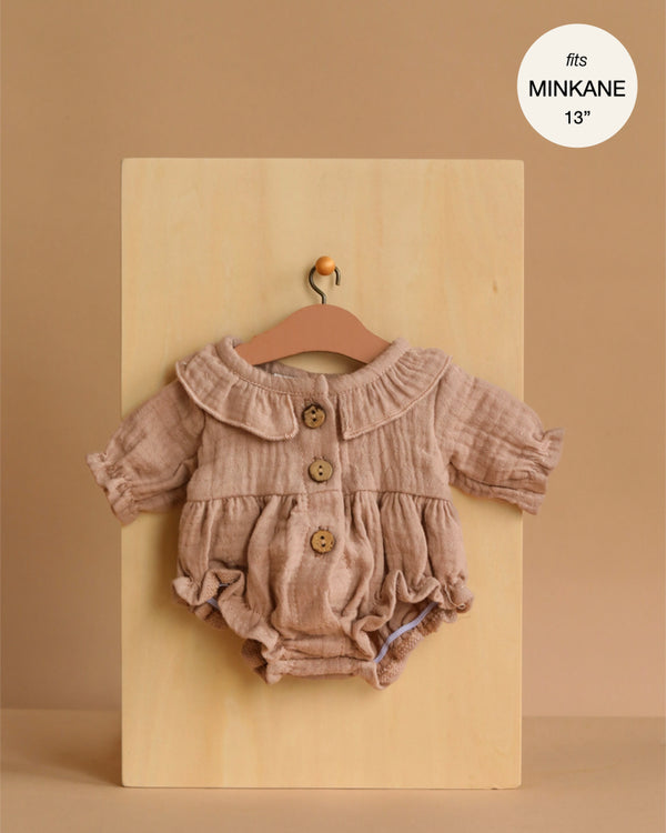The Minikane Doll Clothing | Leonore Ruffled Romper in Cotton Gauze - Latte is a light brown, long-sleeved romper with ruffles and wooden buttons, designed for 13" dolls like Minikane Gordis. Displayed on a hanger against a beige board.