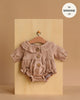 The Minikane Doll Clothing | Leonore Ruffled Romper in Cotton Gauze - Latte is a light brown, long-sleeved romper with ruffles and wooden buttons, designed for 13" dolls like Minikane Gordis. Displayed on a hanger against a beige board.