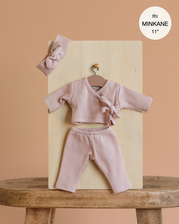 The Minikane 11" Doll Clothing | Raxane Wrap Set & Headband in Ribbed Knit - Petal Pink features a light pink wrap top, matching pants, and a knotted headband displayed on a wooden board against a beige background. Fits Minikane 11” dolls.