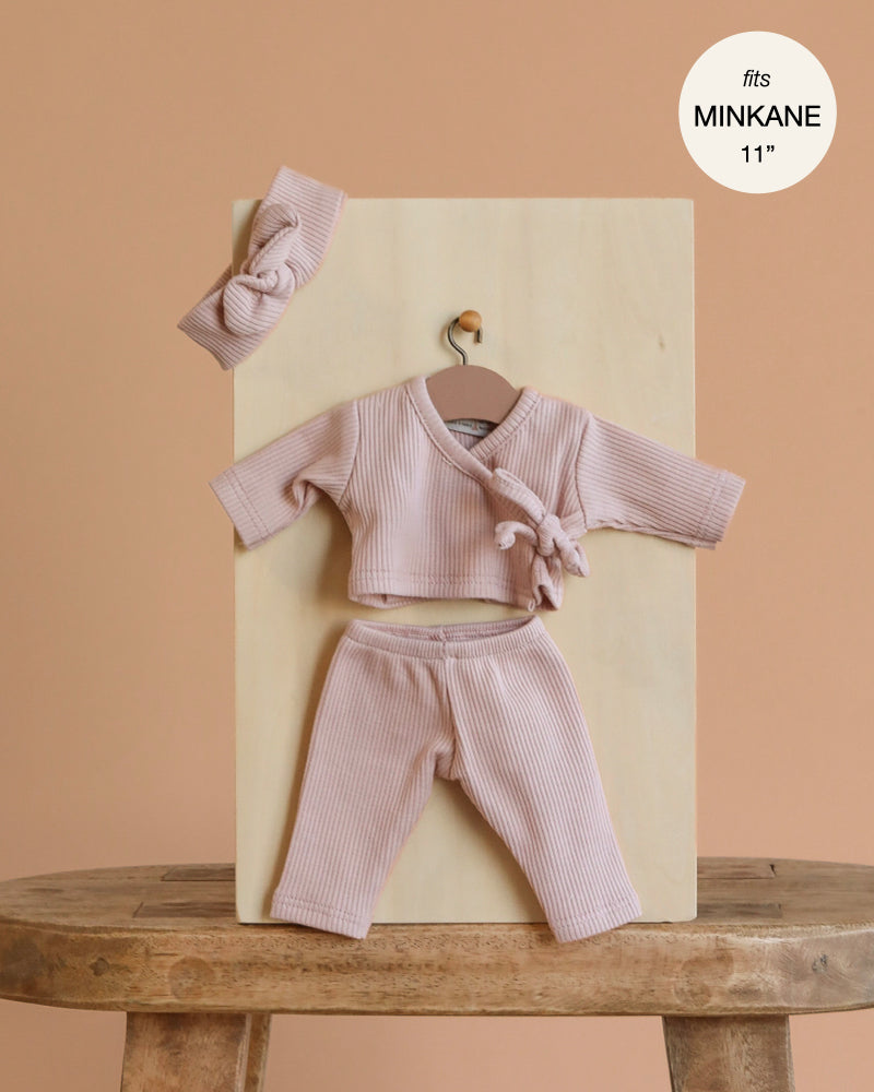 The Minikane 11" Doll Clothing | Raxane Wrap Set & Headband in Ribbed Knit - Petal Pink features a light pink wrap top, matching pants, and a knotted headband displayed on a wooden board against a beige background. Fits Minikane 11” dolls.