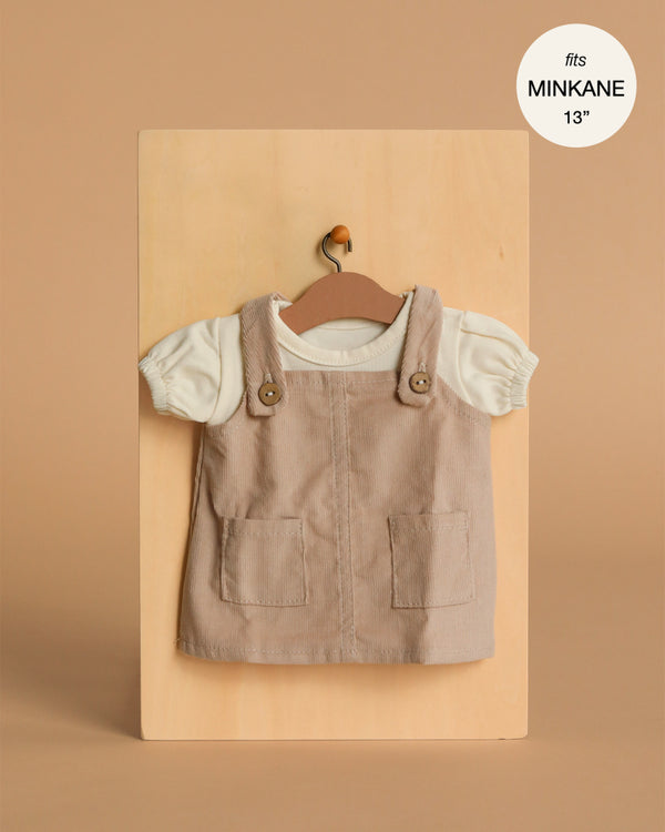 A small beige Minikane Doll Clothing | Nelly Corduroy Dress & T-Shirt set for 13" dolls, with a pinafore featuring two front pockets and a white short-sleeve shirt, displayed on a hanger against a light wooden background.
