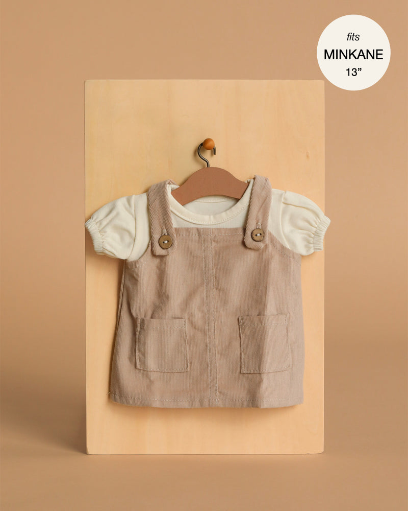 A small beige Minikane Doll Clothing | Nelly Corduroy Dress & T-Shirt set for 13" dolls, with a pinafore featuring two front pockets and a white short-sleeve shirt, displayed on a hanger against a light wooden background.