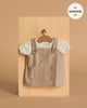 A small beige Minikane Doll Clothing | Nelly Corduroy Dress & T-Shirt set for 13" dolls, with a pinafore featuring two front pockets and a white short-sleeve shirt, displayed on a hanger against a light wooden background.