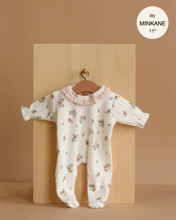 The Minikane 11" Doll Clothing | Camille Onesie in Ribbed Cotton - Pink Flowers features a white ribbed romper with pink ruffle collar and floral print, plus a matching collar and headband. Shown on a wooden hanger, fits MINKANE 11".