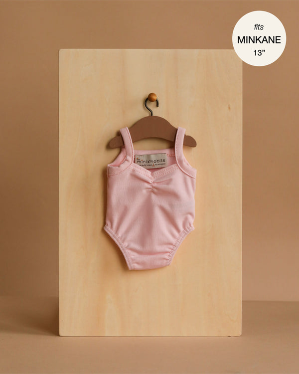 The Minikane 13" Doll Clothing | Anna Leotard in Jersey Cotton - Pale Pink is displayed on a wooden hanger against a light wood board. A circular label in the top right corner reads "fits 13 inch dolls.
