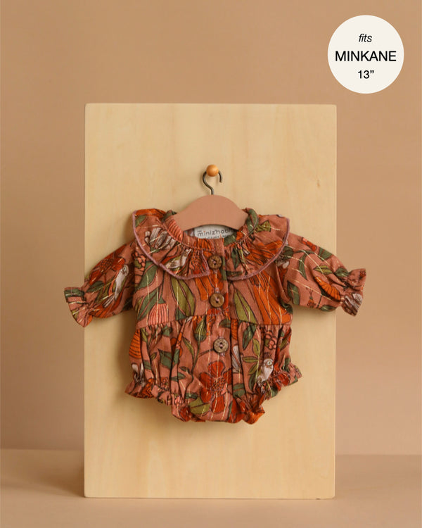 The Minikane 13" Doll Clothing | Leonore Ruffled Romper in Viscose - Birds of the Islands, a floral long-sleeve romper with ruffled collar and sleeves, is shown on a small hanger against a wooden board. Text: "fits Minikane Gordis dolls 34cm".