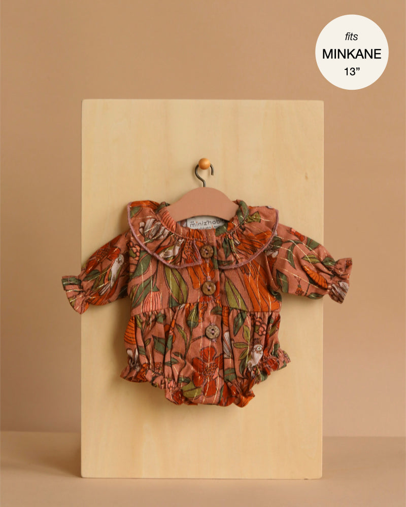 The Minikane 13" Doll Clothing | Leonore Ruffled Romper in Viscose - Birds of the Islands, a floral long-sleeve romper with ruffled collar and sleeves, is shown on a small hanger against a wooden board. Text: "fits Minikane Gordis dolls 34cm".