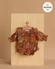 The Minikane 13" Doll Clothing | Leonore Ruffled Romper in Viscose - Birds of the Islands, a floral long-sleeve romper with ruffled collar and sleeves, is shown on a small hanger against a wooden board. Text: "fits Minikane Gordis dolls 34cm".