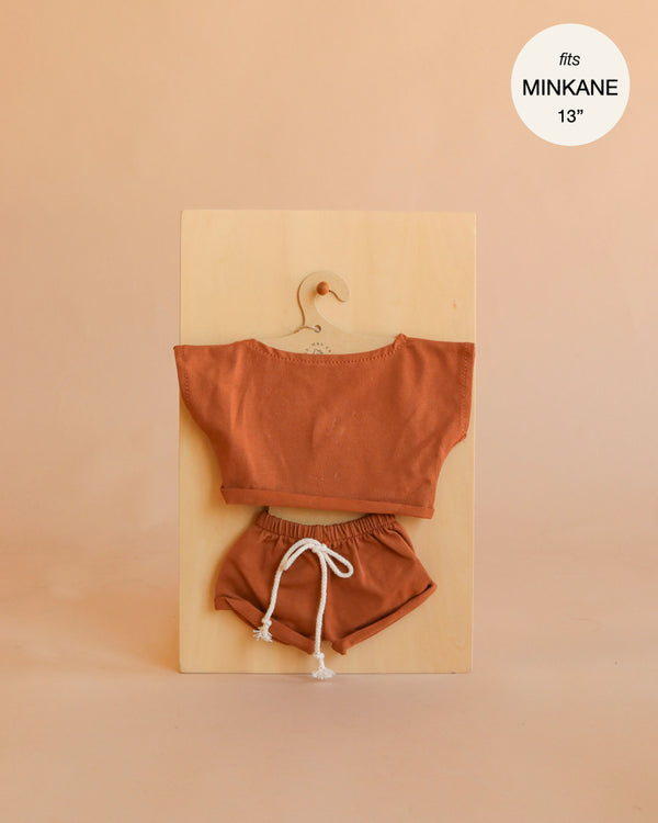 Mrs. Ertha Doll Clothes - 13″-14″ - Rusted Comfy Set, featuring a rust-colored handmade short-sleeve top and drawstring shorts, perfectly sized for Minikane dolls, is displayed on a wooden hanger against a beige background. Text in the corner reads “fits MINIKANE 13”.“.