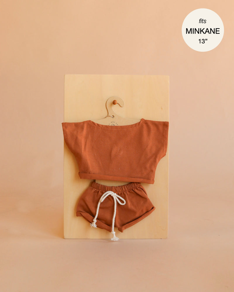 Mrs. Ertha Doll Clothes - 13″-14″ - Rusted Comfy Set, featuring a rust-colored handmade short-sleeve top and drawstring shorts, perfectly sized for Minikane dolls, is displayed on a wooden hanger against a beige background. Text in the corner reads “fits MINIKANE 13”.“.