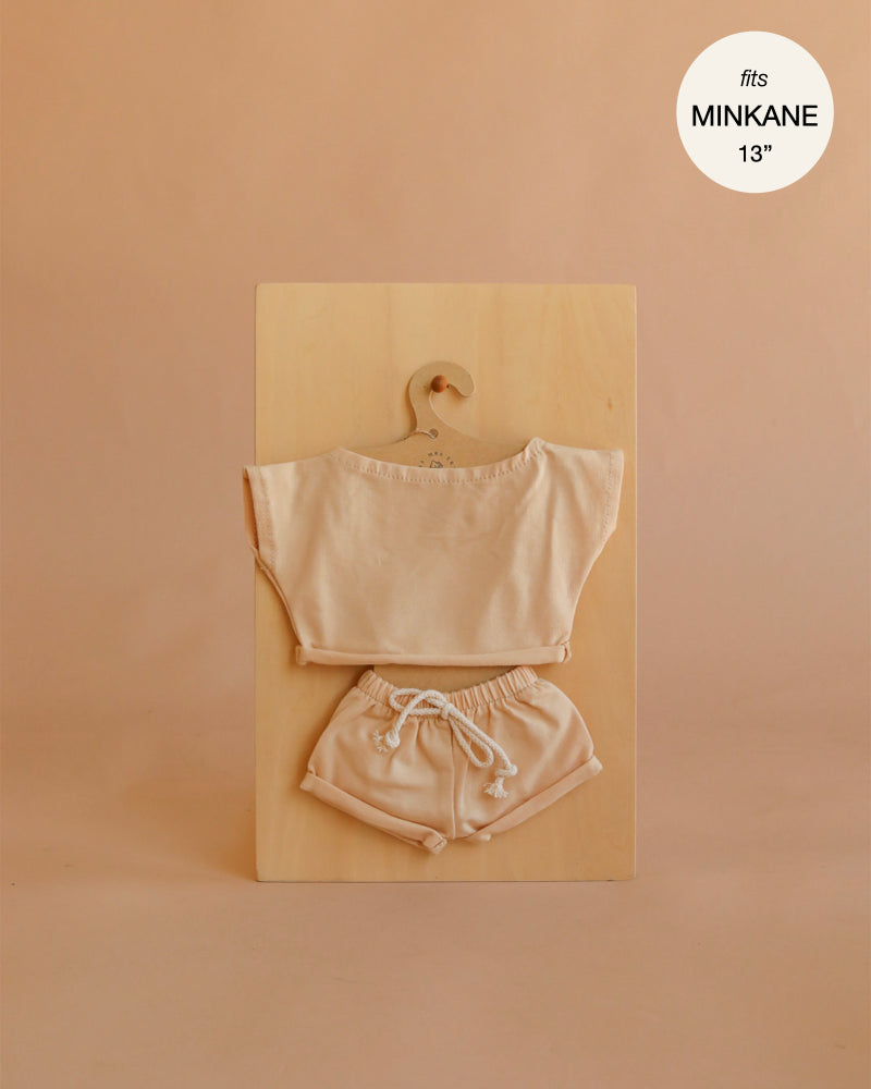 The Mrs. Ertha Doll Clothes - 13″-14″ - Blush Comfy Set, featuring a short-sleeve top and drawstring shorts in a soft beige hue made from organic cotton, is displayed on a wooden hanger against a wooden board. Text in the top right reads “fits Minikane 13” doll clothes.”.