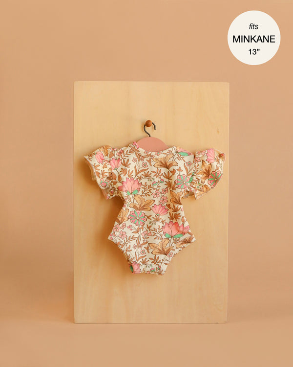 The Minikane 13" Doll Clothing Ondine Bodysuit in Jersey Cotton - Indian Dew, featuring a floral pattern and ruffled sleeves, hangs on a small wooden hanger against a beige background. A label notes it fits Minikane Gordis dolls 13".