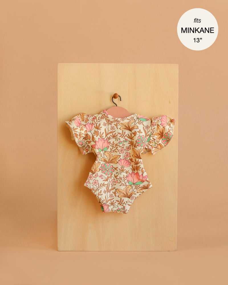 The Minikane 13" Doll Clothing Ondine Bodysuit in Jersey Cotton - Indian Dew, featuring a floral pattern and ruffled sleeves, hangs on a small wooden hanger against a beige background. A label notes it fits Minikane Gordis dolls 13".