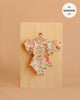 The Minikane 13" Doll Clothing Ondine Bodysuit in Jersey Cotton - Indian Dew, featuring a floral pattern and ruffled sleeves, hangs on a small wooden hanger against a beige background. A label notes it fits Minikane Gordis dolls 13".