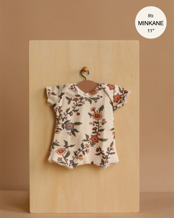 A Minikane 11" Doll Clothing Shorty Bodysuit in Jersey - Gisele hangs on a tiny hanger against light wood. Features short sleeves and a floral pattern. Made in France, fits Minikane Dolls 11". Perfect for dressing your doll in style.