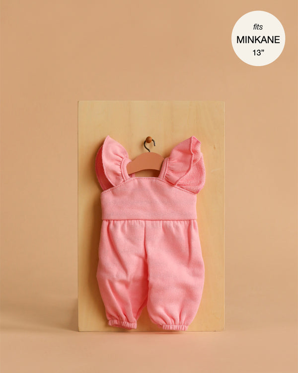 Displayed on a small wooden hanger, the Minikane 13" Doll Clothing | Claudie Overalls in Cotton - Strawberry Pink features ruffled straps and is perfect for 13 inch dolls. The soft pink overalls add charm to any doll clothes collection.