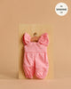 Displayed on a small wooden hanger, the Minikane 13" Doll Clothing | Claudie Overalls in Cotton - Strawberry Pink features ruffled straps and is perfect for 13 inch dolls. The soft pink overalls add charm to any doll clothes collection.