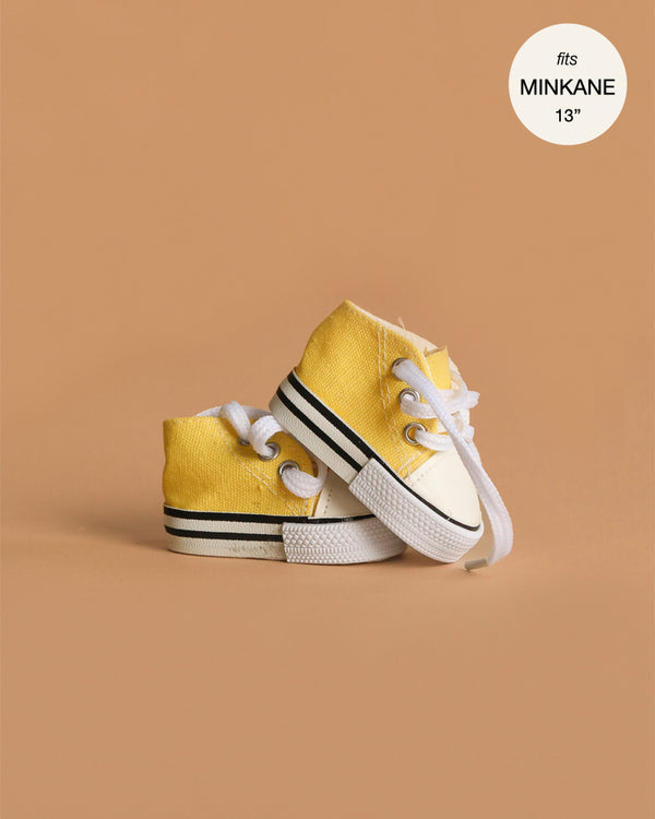 A pair of Minikane Doll Shoes | Converse Style Lemon Sneakers, yellow and white with white laces, for baby dolls, sits on a light brown background. Overlay in the top right reads: "fits Minikane Gordis 34cm".