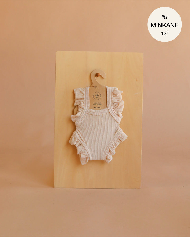 A blush-colored, ribbed handmade bodysuit with ruffled sleeves from the Mrs. Ertha Doll Clothes collection hangs on a small wooden hanger against a light wooden board. Crafted from soft organic cotton and designed to fit 13″-14″ dolls, this charming romper is perfect for your doll’s wardrobe. Text in the corner reads: “fits Minikane 13′′.”.