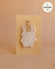 A blush-colored, ribbed handmade bodysuit with ruffled sleeves from the Mrs. Ertha Doll Clothes collection hangs on a small wooden hanger against a light wooden board. Crafted from soft organic cotton and designed to fit 13″-14″ dolls, this charming romper is perfect for your doll’s wardrobe. Text in the corner reads: “fits Minikane 13′′.”.