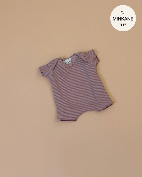 Displayed on a light brown background, the Minikane 11" Doll Clothing | Dark Orchid Ribbed Body Shorty is a small, short-sleeved onesie in mauve. The top right corner sports a circular tag reading "fits Minikane 11". This knit piece showcases exquisite French craftsmanship.