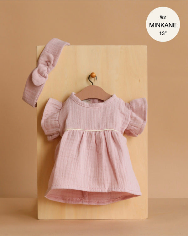 The Minikane Doll Clothing | Daisy Dress & Headband in Cotton Gauze - Orchid Pink is a pale pink dress with puff sleeves and a matching headband, displayed on a wooden board. Text reads "fits Minikane 13 inch dolls." Background is warm beige.