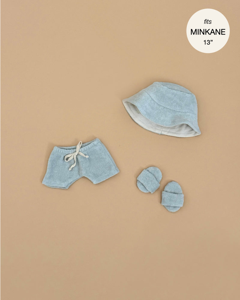 Green Tea terrycloth 3-piece swimsuit set by Emile & Ida for 13-inch Minikane dolls, featuring shorts, a bucket hat, and matching booties, styled on a beige background.