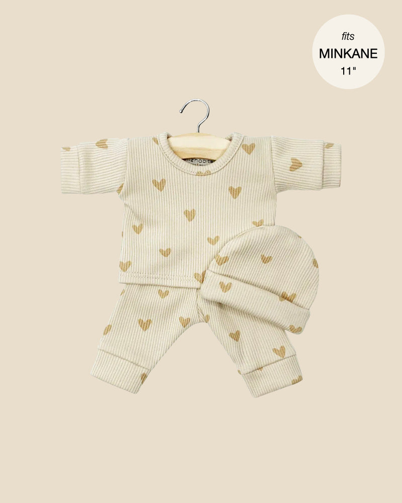 The Minikane Doll Clothing set, "Heart Print Pajamas," includes a beige, ribbed long-sleeve top, pants, and beanie with heart patterns. Displayed on a small hanger against a beige backdrop, this handmade French outfit fits MINKANE 11-inch dolls perfectly.