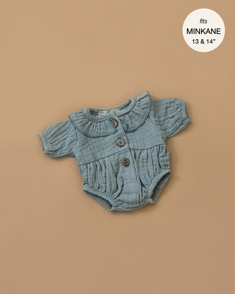 A small, textured Leonore ruffled jumpsuit in Baltic Green with short sleeves and a Peter Pan collar, adorned with three buttons down the front. Specially designed for Minikane dolls, it is displayed on a beige background. A circular label in the corner states "fits Minikane 13 & 14".