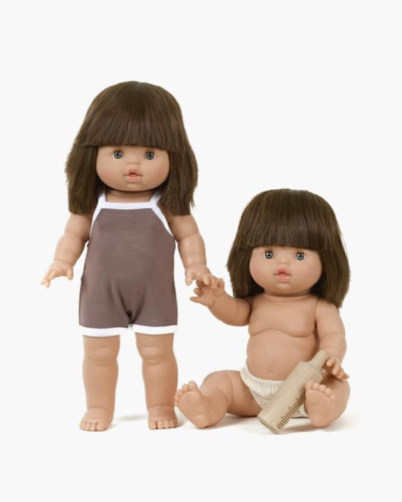 Two Minikane Chléa dolls with short brown hair feature similar expressions. One stands in a brown sleeveless outfit, while the other sits in a diaper holding a comb. Both exude a subtle vanilla scent for added charm.