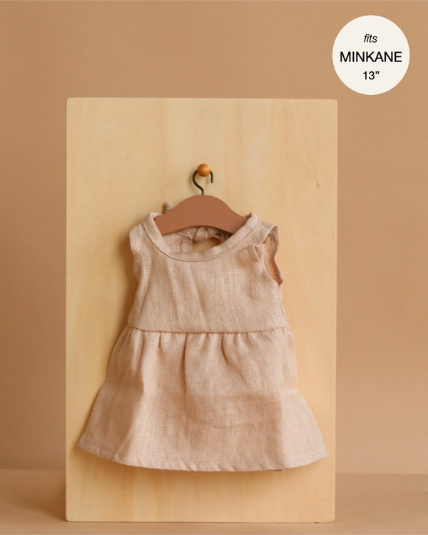 The Minikane 13" Doll Sleeveless Faustine Dress in Washed Linen - Nude, featuring a gathered skirt, is shown on a wooden hanger against a board. Text reads “fits 13 inch doll.” A quality addition to your doll clothing collection.