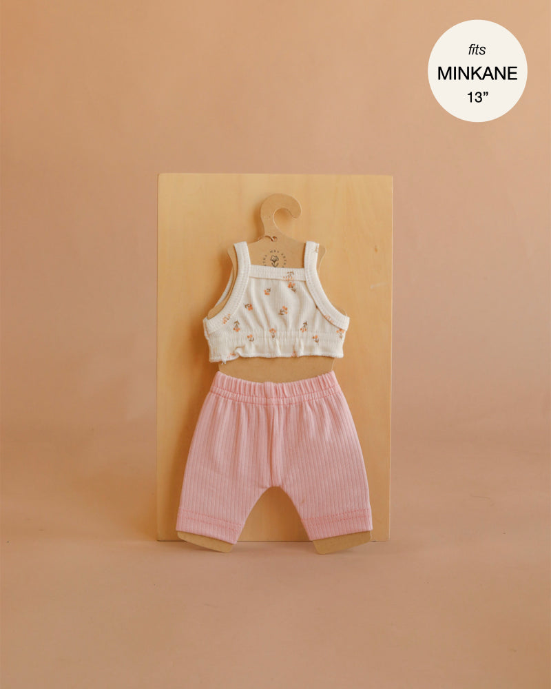 A charming small doll outfit, the Mrs. Ertha Doll Clothes - 13″-14″ - Flower Buds Leggings Set features a handmade white floral crop top paired with pink ribbed pants, both crafted from soft organic cotton. Neatly displayed on a hanger against a beige background, this set is designed to fit Minikane dolls sized 13”-14”.