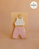 A charming small doll outfit, the Mrs. Ertha Doll Clothes - 13″-14″ - Flower Buds Leggings Set features a handmade white floral crop top paired with pink ribbed pants, both crafted from soft organic cotton. Neatly displayed on a hanger against a beige background, this set is designed to fit Minikane dolls sized 13”-14”.