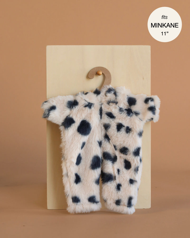 The Minikane 11" Doll Clothing | Little Cat Jumpsuit in Faux Fur, a small white furry jumpsuit with black spots, is shown on a wooden hanger against a beige backdrop. Fits MINIKANE 11'' dolls and adds charm to your collection.