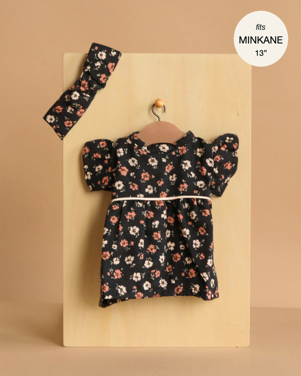 The Minikane Doll Clothing | Rylee+Cru | Daisy Dress and Headband - Capucine, a black floral puff-sleeve dress with a white waist trim for 13” and 14” dolls, hangs on a small hanger with its matching headband on a light wooden board.