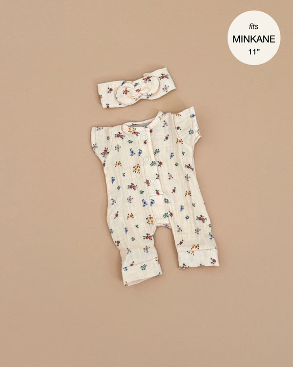 The Lili Jumpsuit in Elise Cotton Gauze, a cream floral-patterned doll outfit complete with buttons and matching headband, is showcased on a beige backdrop. Designed for Minikane dolls, it features the label "fits MINKANE 11," and is made in France. Hand washable for easy care.