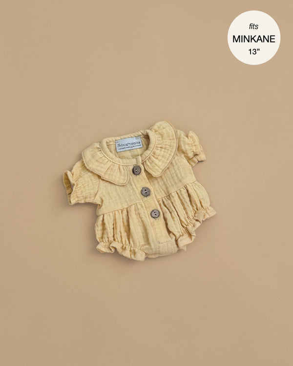 A light yellow Leonore Ruffled Romper in vanilla cotton gauze, featuring a Peter Pan collar, ruffled hem, and three brown buttons—perfectly sized for Minikane Gordis dolls. Text reads: “fits MINIKANE 13''.”.