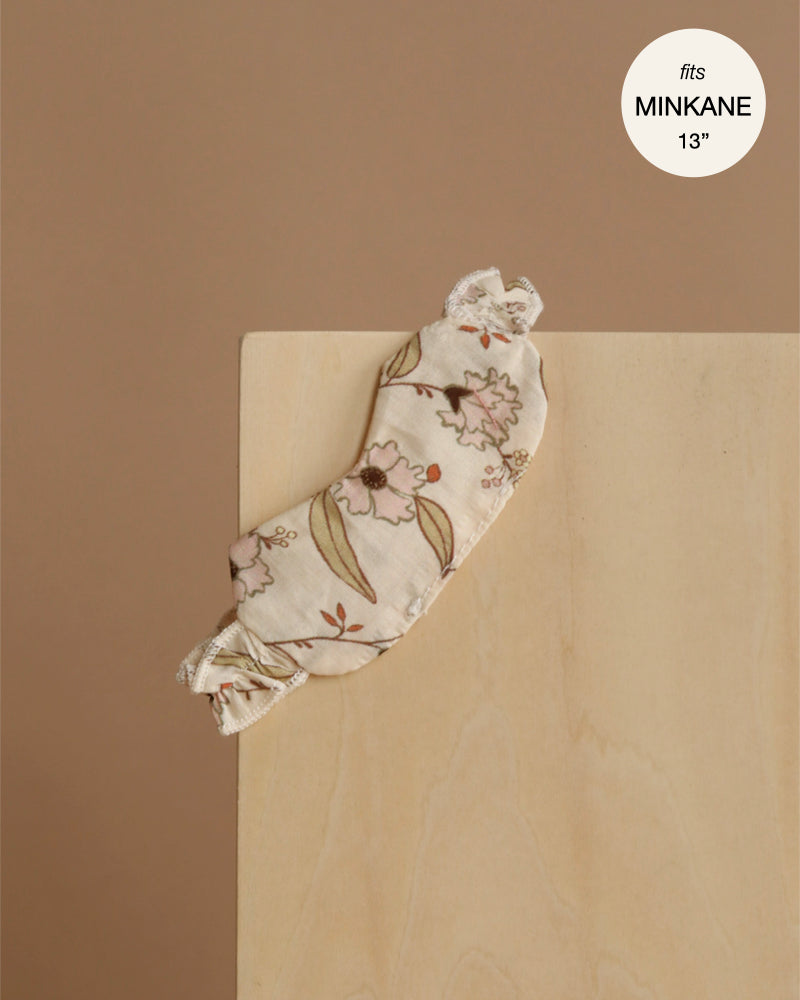 The Minikane Doll Clothing | Janelle Sleep Mask in Ruffled Cotton, featuring floral fabric and lace trim, rests on light wood against a beige background. Fits 13 inch dolls—perfect for adding charm to any doll clothes collection. Label reads "fits MINKANE 13.