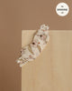 The Minikane Doll Clothing | Janelle Sleep Mask in Ruffled Cotton, featuring floral fabric and lace trim, rests on light wood against a beige background. Fits 13 inch dolls—perfect for adding charm to any doll clothes collection. Label reads "fits MINKANE 13.