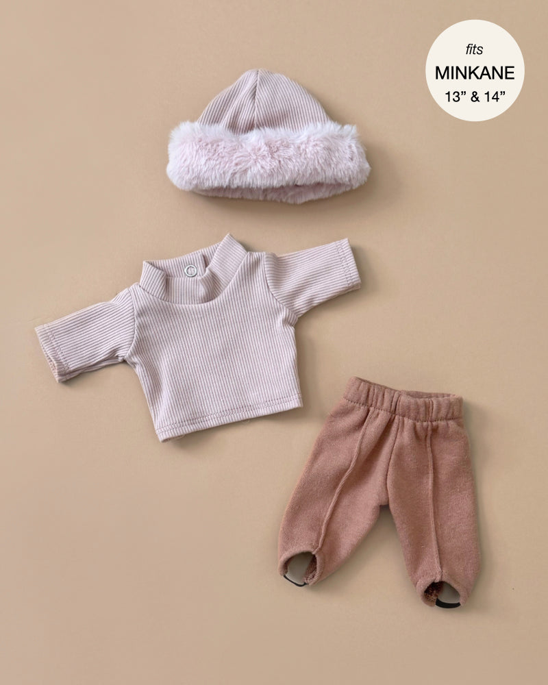 The Minikane Doll Clothing set features a ribbed pink long-sleeve sweater, pink pants with elastic cuffs, and a pink and white fluffy hat. Designed to fit 13” & 14” Minikane dolls, these French-crafted pieces emphasize elegance on a beige backdrop.