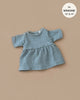 The Faustine dress in Baltic Green, designed for Minikane dolls 13" and 14", is beautifully textured with short sleeves and showcased against a beige background.