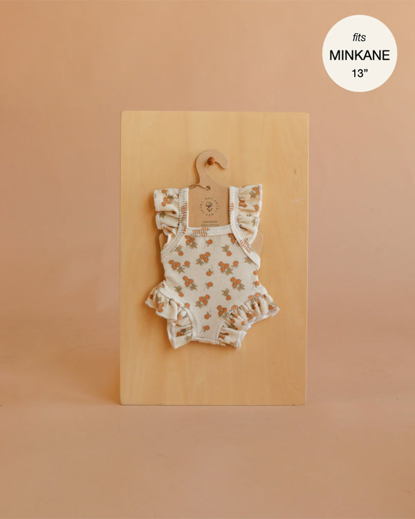 Displayed on a wooden board with a hanger, the Mrs. Ertha Doll Clothes - 13″-14″ Raspberry Bunches Frilled Bodysuit showcases charming floral ruffles, making it a delightful addition to any Minikane doll’s wardrobe. A label in the top right corner indicates “fits MINKANE 13”.