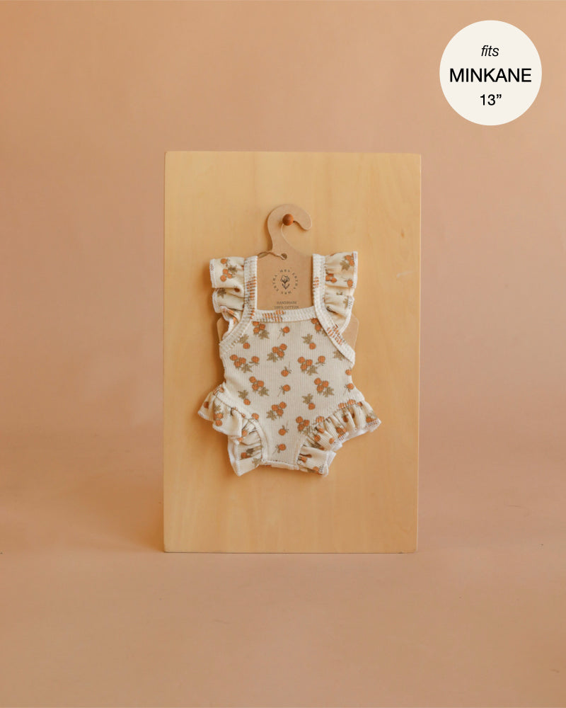 Displayed on a wooden board with a hanger, the Mrs. Ertha Doll Clothes - 13″-14″ Raspberry Bunches Frilled Bodysuit showcases charming floral ruffles, making it a delightful addition to any Minikane doll’s wardrobe. A label in the top right corner indicates “fits MINKANE 13”.