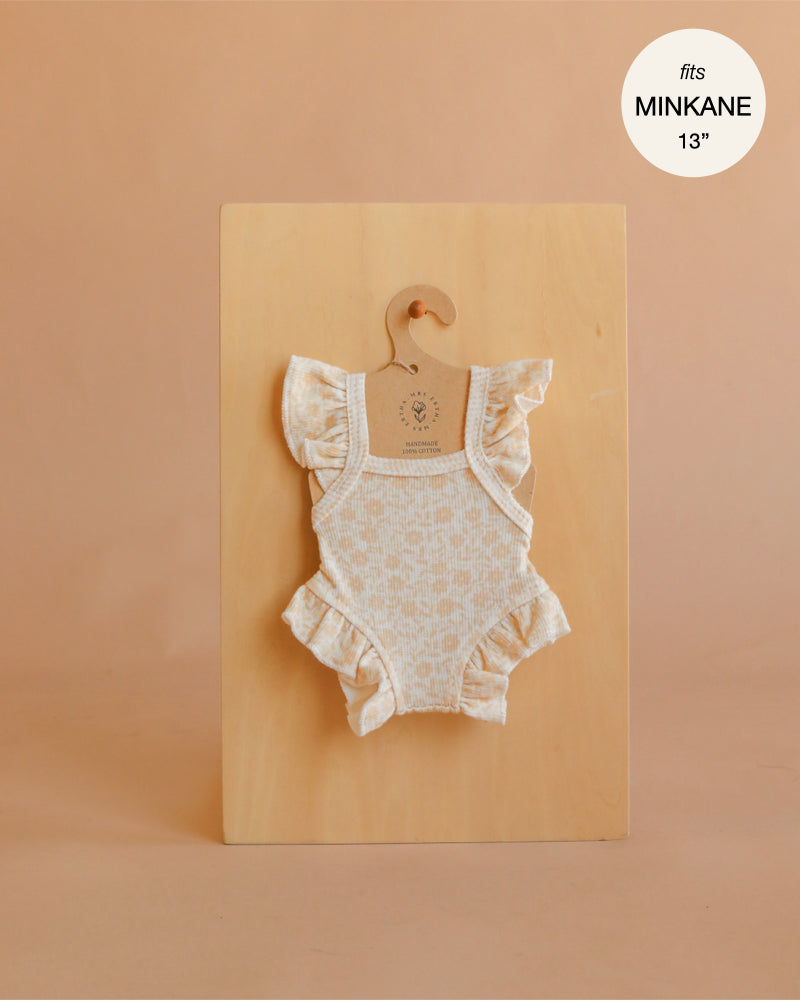 A cream-colored Mrs. Ertha Doll Clothes Bouquet Silhouette Frilled Bodysuit, designed for 13″-14″ dolls, hangs on a wooden hanger against a beige background. Featuring delicate ruffles and a subtle floral pattern, this organic cotton ensemble is the perfect addition to any Minikane doll clothes collection. A circle in the corner reads “fits MINIKANE 13”.”.