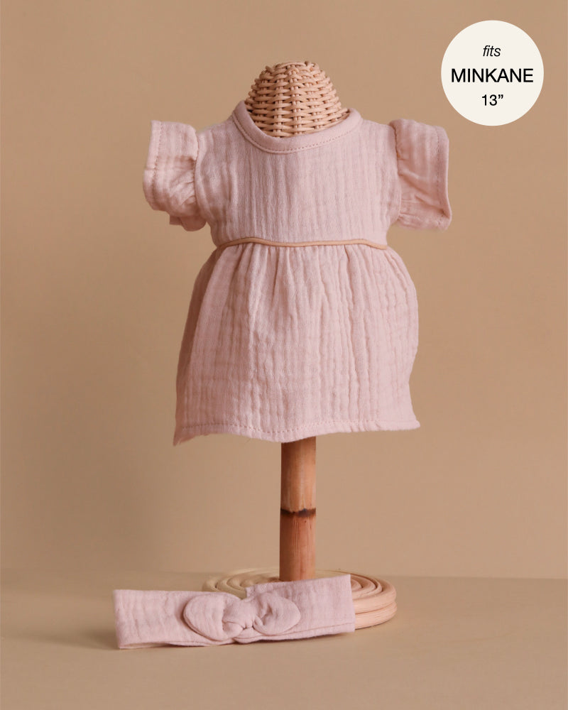 The Minikane Doll Clothing Daisy Dress & Headband in Cotton Gauze - Orchid Pink is a light pink, short-sleeved set for 13" Minikane dolls and also fits as 14" doll clothes, shown on a wicker mannequin stand.