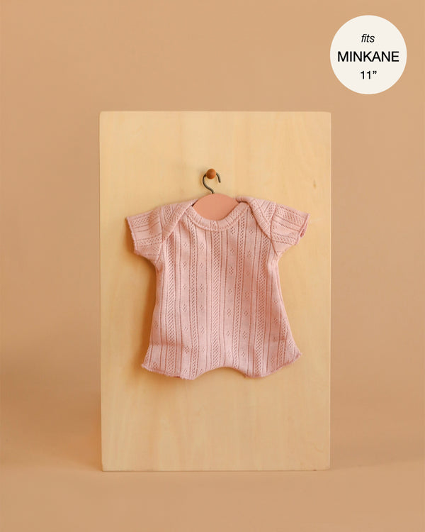 A light pink, short-sleeved dotted knit bodysuit for Minikane 11" dolls hangs on a small hanger against a wooden board. Beige background highlights this charming "Minikane 11\" Doll Clothing | Shorty Bodysuit in Dotted Knit - Petal".
