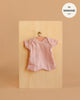 A light pink, short-sleeved dotted knit bodysuit for Minikane 11" dolls hangs on a small hanger against a wooden board. Beige background highlights this charming "Minikane 11\" Doll Clothing | Shorty Bodysuit in Dotted Knit - Petal".