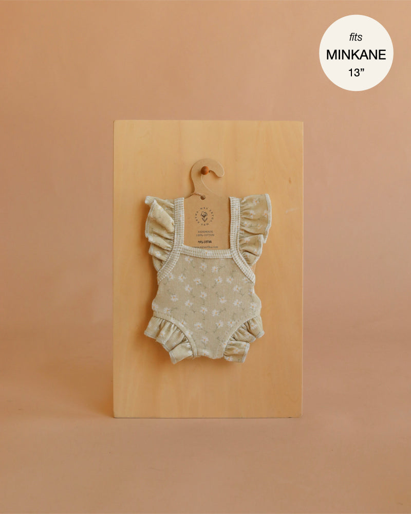A Mrs. Ertha Doll Clothes - 13″-14″ - Little Daisys Frilled Bodysuit, featuring delicate white floral patterns and frilled details in beige, hangs on a small hanger against a wooden board. Ideal for dressing Minikane dolls, it includes a label stating “fits MINKANE 13”.